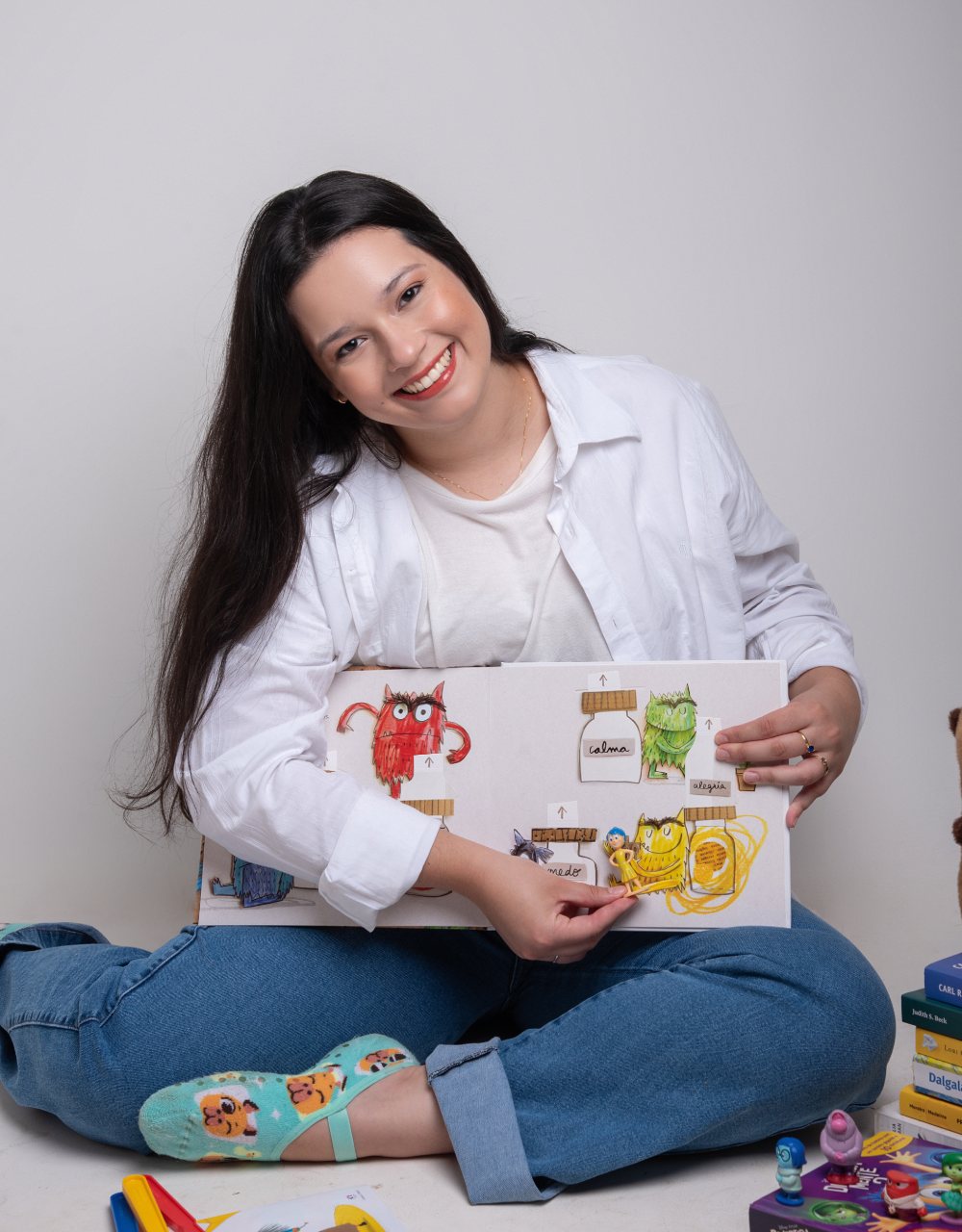 Karine sentada no chão mostrando um livro infantil sobre emoções, com brinquedos ao redor